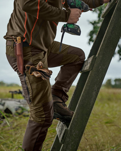 Willow Green Deep Brown Coloured Harkila Scandinavian Trousers on forest background #colour_willow-green-deep-brown
