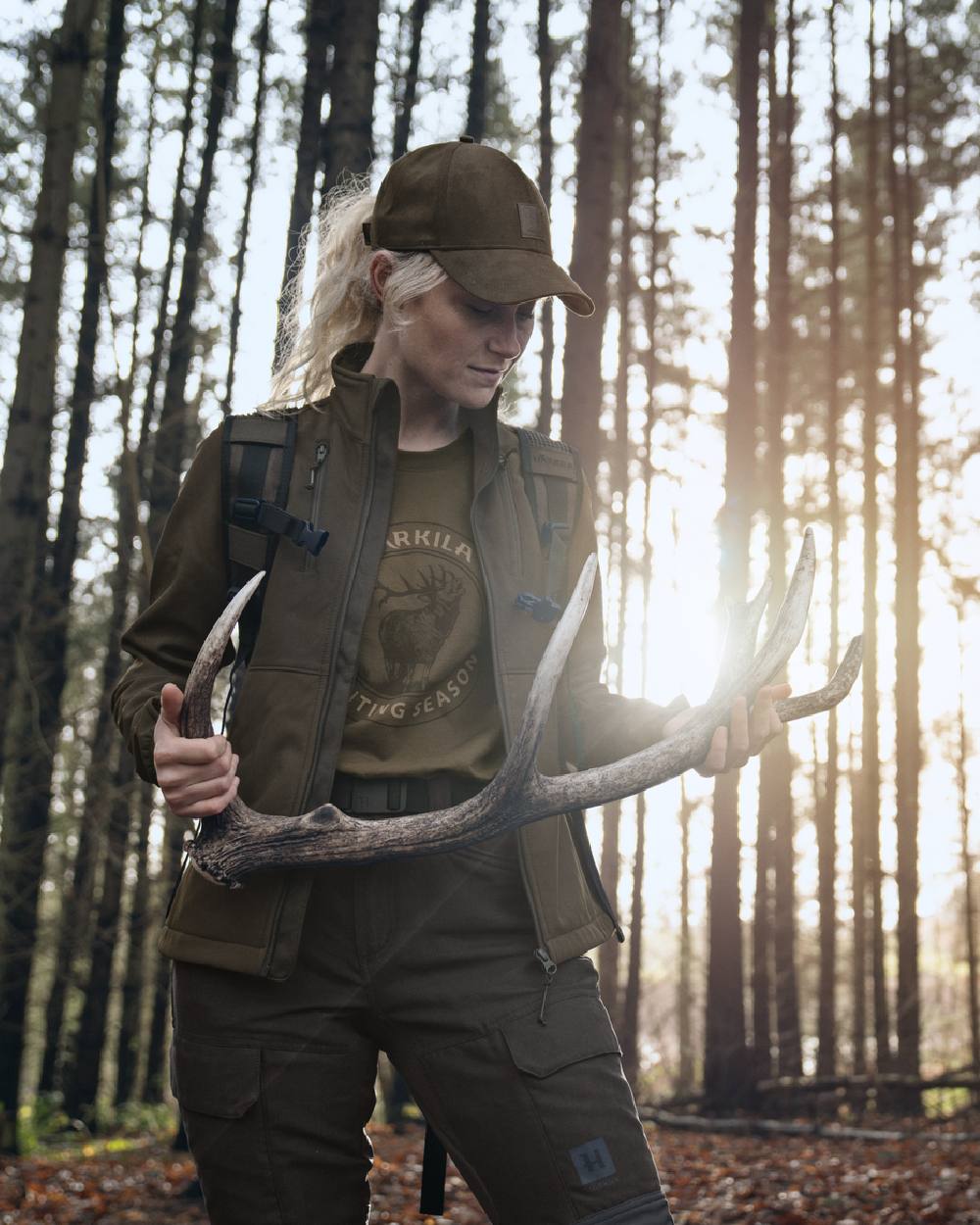 A woman in outdoor hunting apparel stands in a sunlit forest holding a large deer antler. She wears a brown cap, olive green vest, dark olive shirt with a graphic print, and dark cargo pants. #colour_beech