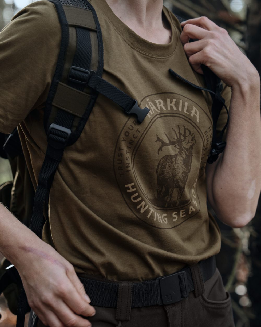 A close-up of a person wearing a brownish-olive tactical t-shirt with a dark brown circular seal graphic featuring an elk and the text 'ARKILA HUNTING SEASON'. The person is also wearing a black backpack and dark pants. #colour_beech