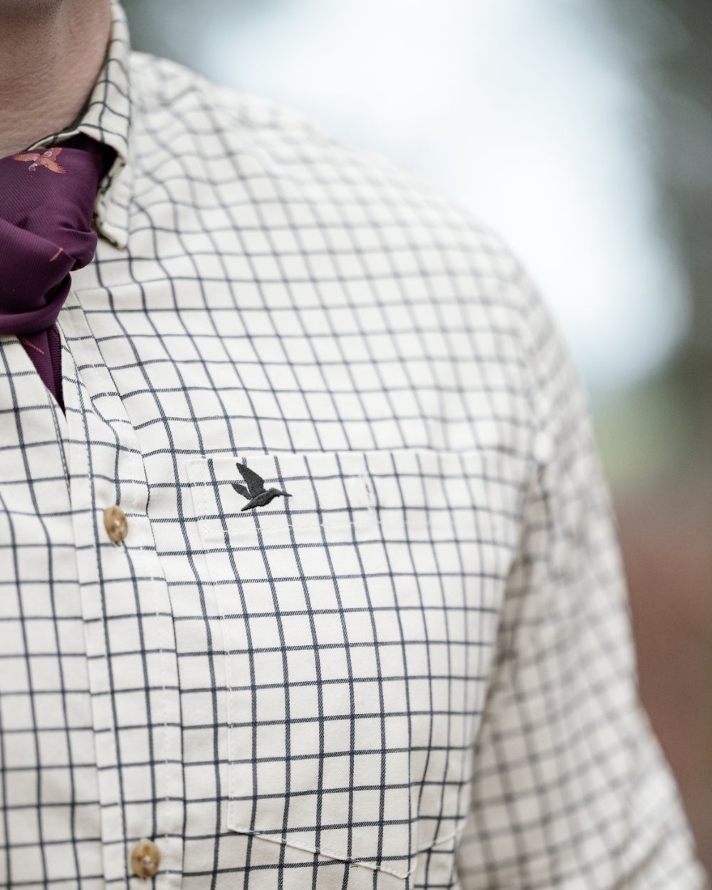 Close-up of a person wearing a white button-down shirt with a dark blue grid pattern, a dark purple necktie, and a small embroidered bird logo on the chest pocket. #colour_dark-sapphire-check