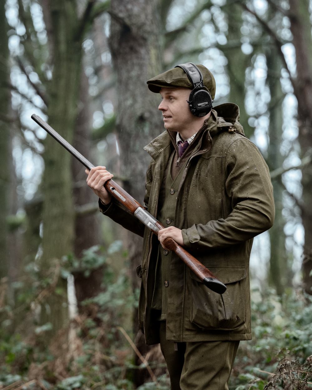 A man in a forest wearing a brown deerstalker hat, over-ear headphones, and an olive green hunting jacket, holding a shotgun. #colour_dark-sapphire-check