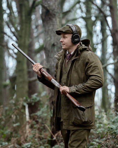 A man in a forest wearing a brown deerstalker hat, over-ear headphones, and an olive green hunting jacket, holding a shotgun. #colour_dark-sapphire-check
