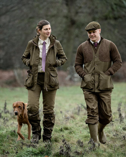 A man and a woman wearing matching brown hunting suits walk in a grassy field with a brown dog. The man wears a flat cap, and both wear olive green jackets, matching trousers, and rubber boots. #colour_estate-blue-pine-green-check